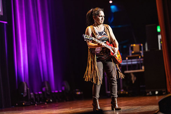 Mott, Motown and More event photo of woman holding a right handed electric guitar dressed in high heeled boots and a fringed vest